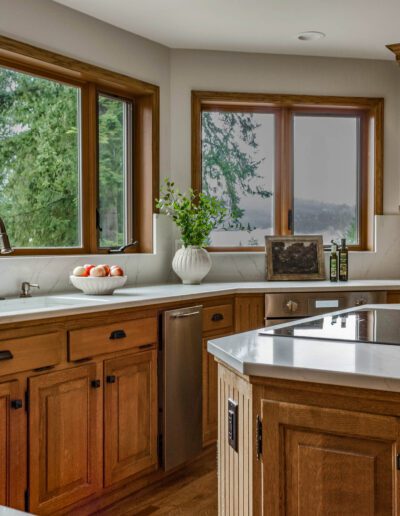 Modern kitchen interior with wooden cabinets, stainless steel appliances, and a large island, overlooking a forest through expansive windows.