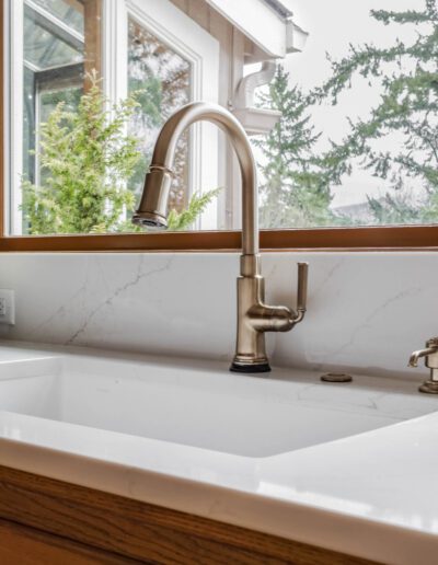 A bright, modern kitchen sink with stainless steel faucet, surrounded by greenery visible through large window, and a bowl of apples on the counter.