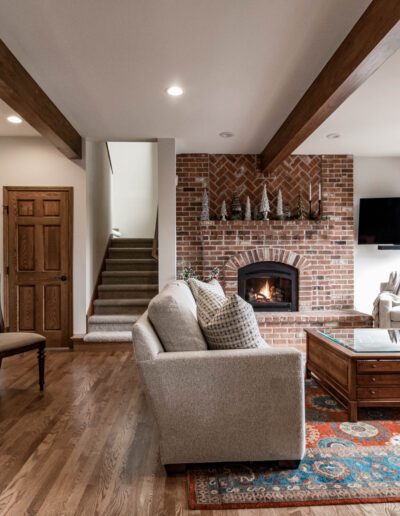 A cozy living room with exposed brick fireplace, wooden beams on the ceiling, hardwood floors, and elegant furnishings, leading to a sunny patio.