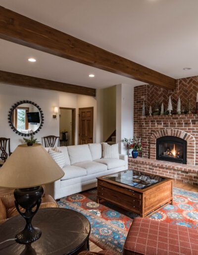 Cozy living room with a brick fireplace, white sofas, wooden ceiling beams, and a shelf full of decorative items.