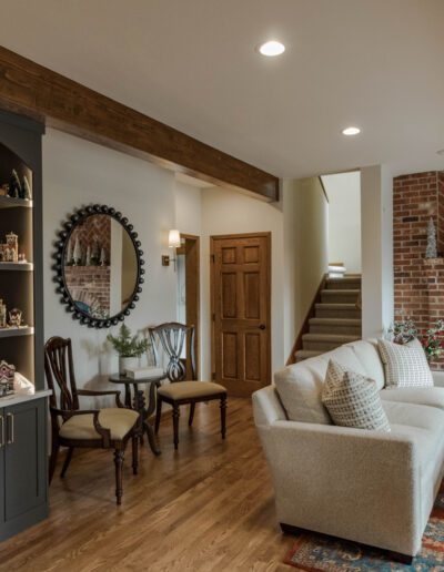 Cozy living room decorated for christmas with a brick fireplace, festive village display on a cabinet, and comfortable seating areas.