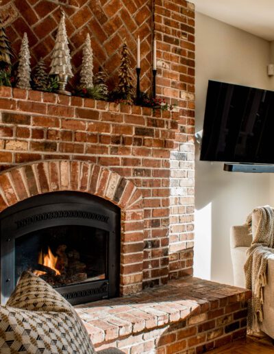 Cozy living room with a lit fireplace, brick walls, a mounted tv, and comfortable armchairs, decorated with festive garlands.