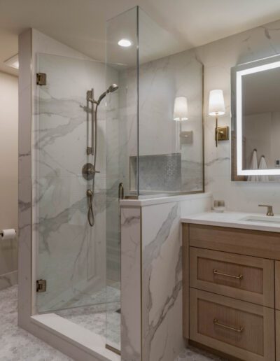 A modern bathroom with a glass-walled shower, marble walls, a wooden vanity with a basin, and a large illuminated mirror.