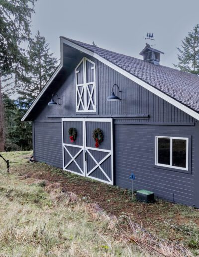 A gray barn-style house with a steep pitched roof and wreaths on its double doors, surrounded by tall trees and overgrown grass.