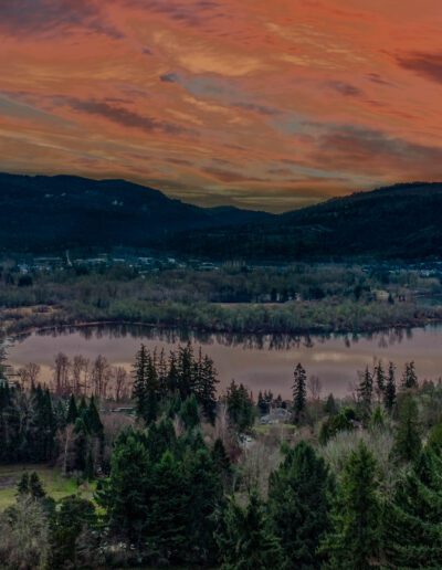 Aerial view of a tranquil lake surrounded by a forest with a village nearby, under a vibrant sunset sky.