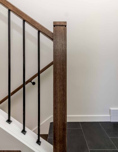 Close-up of a staircase with dark wooden steps, black metal balusters, and a wooden handrail on a tiled floor in a well-lit space with white walls.