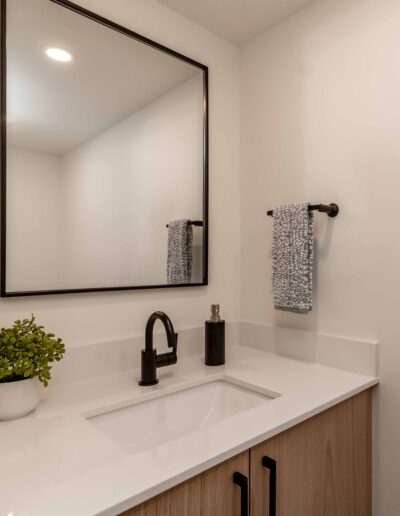 A modern bathroom features a rectangular mirror, a black faucet, a white countertop, a potted plant, and a hand towel on a black holder.