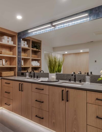 Modern bathroom with a large mirror, dual sinks, wooden cabinets, and open shelving. The shelves are stocked with towels, baskets, and toiletries. A plant and a flower arrangement decorate the counter.