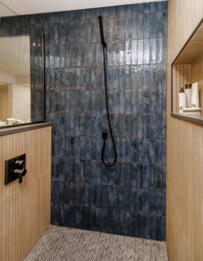 Modern bathroom with a walk-in shower featuring dark blue tiles, a black handheld showerhead, and wood-effect wall panels. The shower area includes built-in shelves with neatly arranged toiletries.