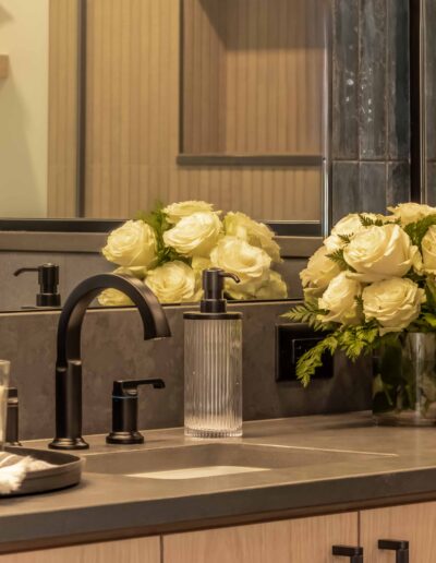 A bathroom sink with black fixtures, a soap dispenser, a candle, a vase of white roses, and folded towels. There is a large mirror reflecting the decor.